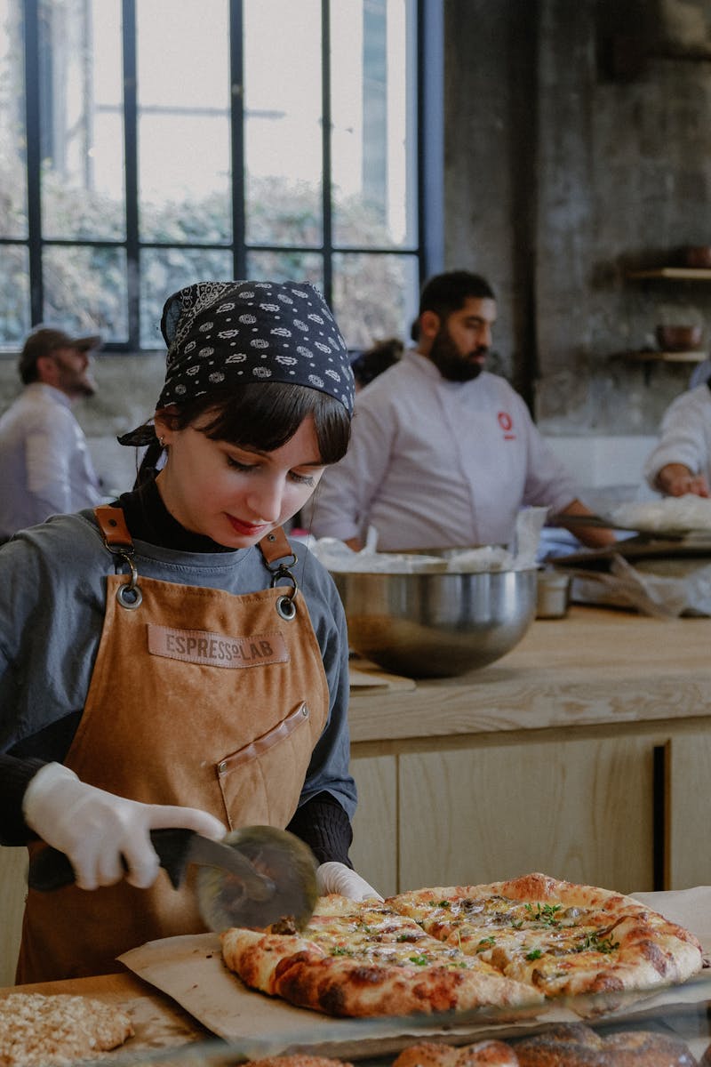 Young female chef slicing pizza in bustling bakery kitchen with breads and coworkers.